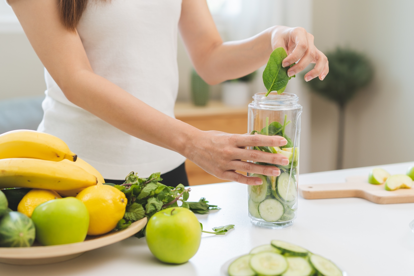 Žena připravuje v kuchyni na lince detoxikační smoothie z okurky, banánu, špenátu a jablka.